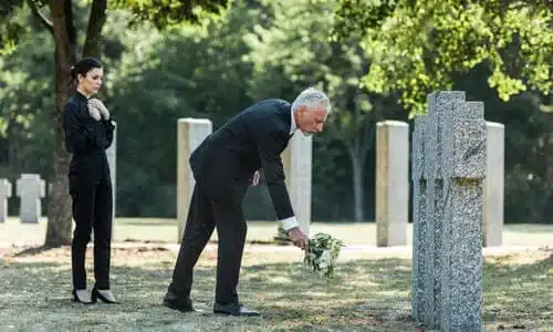An old man leaving a bouquet of flowers at a grave, and a woman mourning behind him.