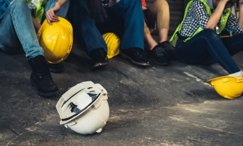 Group of construction workers sitting on the ground with safety helmets scattered, symbolizing a construction accident or workplace injury scene.