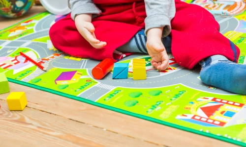 A small child sitting on a colorful playmat surrounded by wooden blocks and toys, representing a potential daycare accident environment.