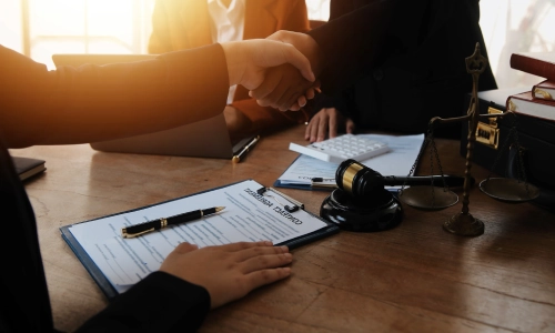 Truck accident legal consultation scene showing attorneys and clients shaking hands at a desk with legal documents, a gavel, and scales of justice.