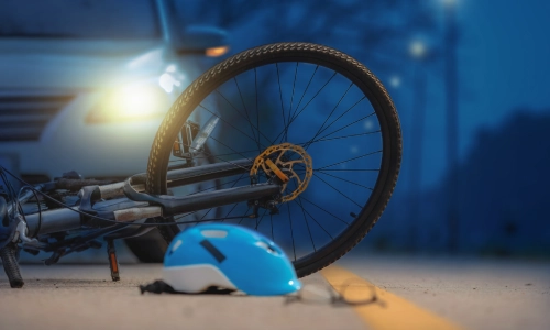 Bicycle accident scene at night showing a damaged bike lying on the road in front of a car alongside a blue helmet and a pair of glasses on the pavement