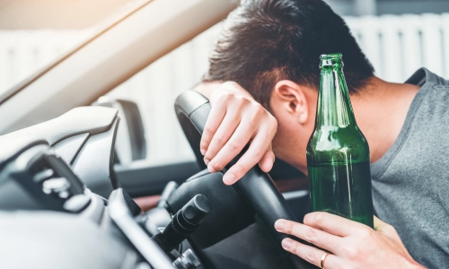 Man slumped over a car steering wheel holding a green beer bottle, symbolizing drunk driving and impaired judgment behind the wheel.