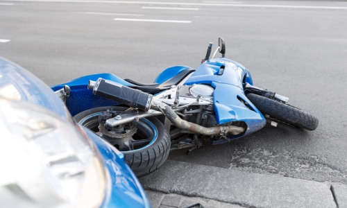 Motorcycle accident involving a blue sports bike lying on its side in front of a car on a city street, showing visible damage to the motorcycle's frame.