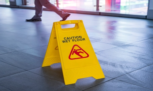 Yellow caution sign reading "Caution Wet Floor" placed on a shiny, wet tile surface in a commercial building, representing a slip and fall accident hazard.