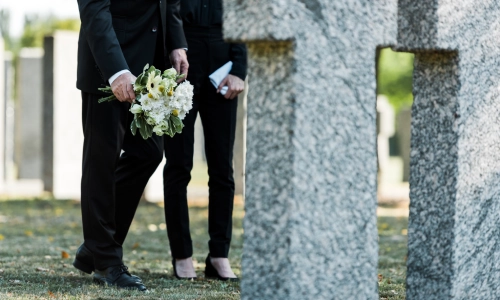 Wrongful death concept shown by two people in formal black attire standing at a cemetery, holding a bouquet of white flowers beside a gravestone.