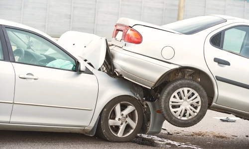 Rear-end car accident showing a silver sedan partially mounted on another vehicle, both with visible damage, airbags deployed, and debris on the road.