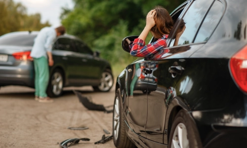 Car accident scene with a distressed woman in a black vehicle and a man inspecting damage to a gray car, with broken car parts scattered on the road.