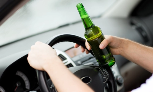 A person holding a green beer bottle while driving a car, with both hands on the steering wheel — representing a Drunk Driving Accident.
