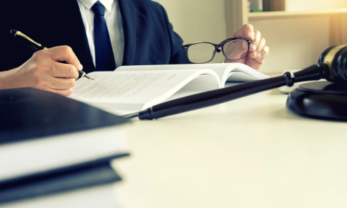 A lawyer sits at a desk, holding glasses and writing notes in a legal book, with a gavel in the foreground — involved in a hit and run accident lawyer case.