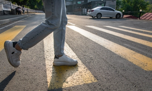 A person walking across a marked pedestrian crosswalk in daylight with a white car parked nearby — involved in a Pedestrian Accident.
