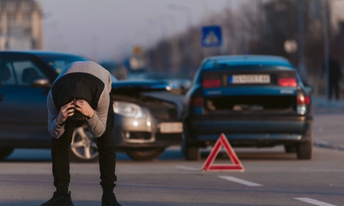 A man bends over holding his head in front of two damaged cars in the middle of a street, with a warning triangle placed nearby, after an uninsured motorist accident.