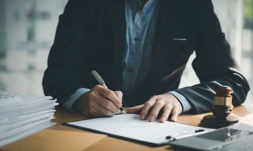 A lawyer is seated at a desk, reviewing and signing legal documents related to a workers' compensation case, with a gavel, tablet, and stacks of paperwork.