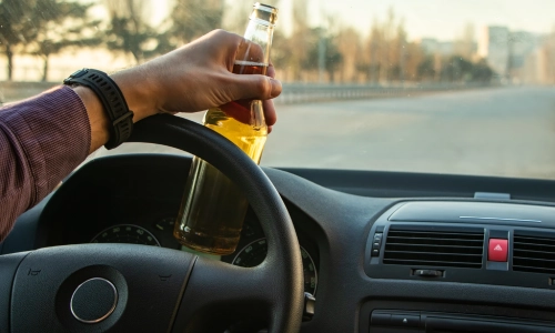 Driver holding a beer bottle while driving on an empty road, depicting a drunk driving accident risk.
