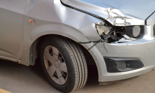 Close-up of a silver car with visible front-end damage from an accident, including a dented hood, broken headlight, and scratched bumper.