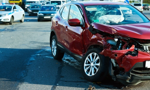 Car accident scene featuring a red SUV with severe front-end damage and a shattered windshield, stopped in the middle of a busy intersection with other vehicles and police presence in the background.
