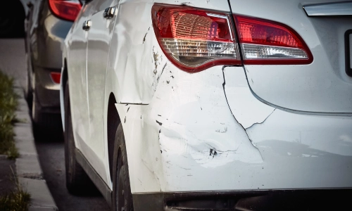 Car accident damage shown on the rear side of a white vehicle with visible scratches, dents, and cracks, parked on the side of the street.