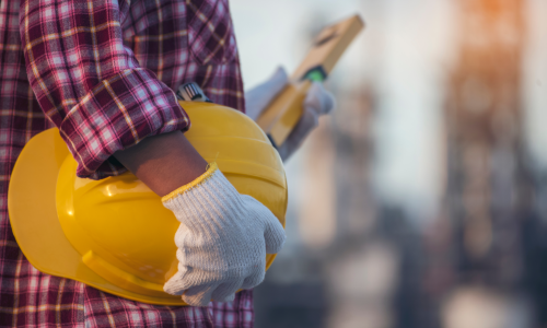 A construction worker in a plaid shirt holding a yellow safety helmet under his arm.