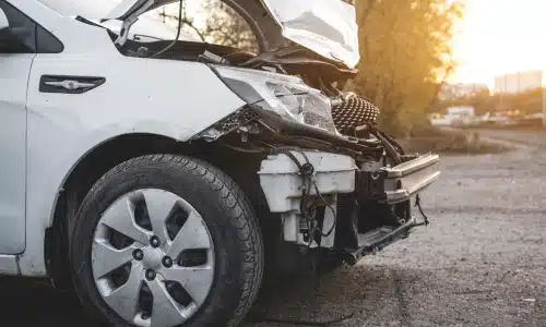 Close-up of a white car with severe front-end damage after a car accident, parked on a dirt roadside at sunset.