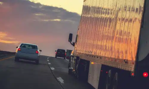 Large commercial truck driving on a highway during sunset with cars ahead on the road.