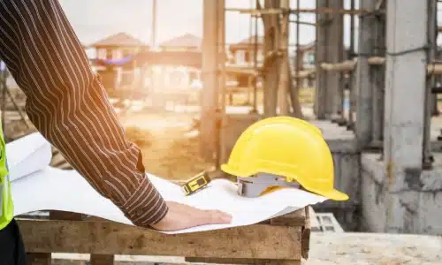 Construction worker reviewing blueprints at a building site with a yellow hard hat and tape measure.