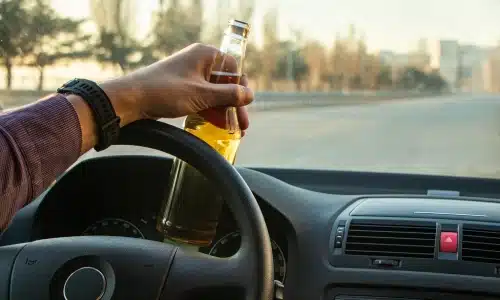 Driver holding a beer bottle behind the wheel on an open road, illustrating the dangerous and illegal behavior leading to a drunk driving accident