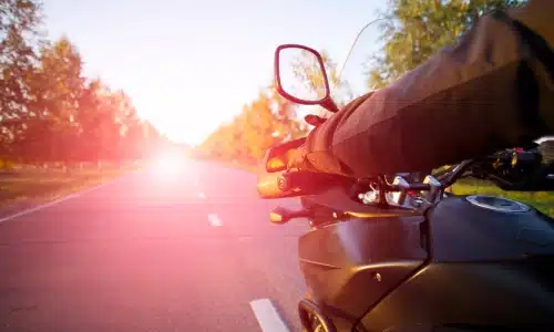 Motorcyclist riding on a scenic road at sunset with visible sun glare and tree-lined sides.
