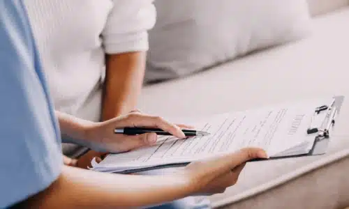 A healthcare worker in scrubs holding a clipboard and pen while speaking with an elderly woman, representing a situation that may involve nursing home abuse