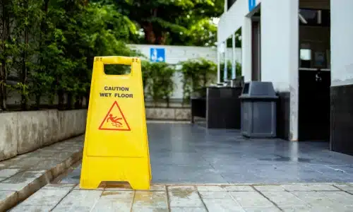 A bright yellow “Caution Wet Floor” sign placed on a slick tile walkway outside a building, warning of a potential slip and fall accident hazard