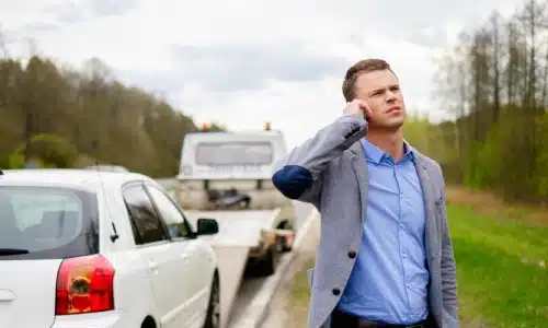 Man standing beside a damaged white car on the roadside, making a phone call after an accident.