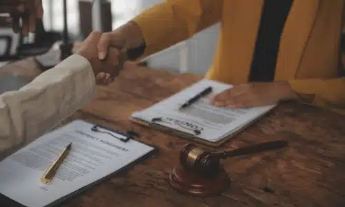Two people shaking hands over contract documents with a gavel on the desk, symbolizing legal agreement.