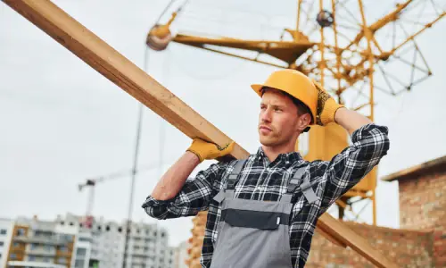 A construction worker holding his helmet and carrying a wooden beam on his shoulder.
