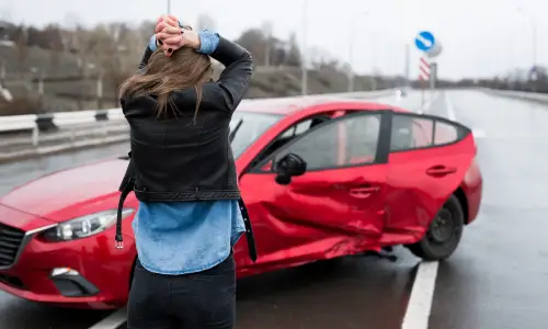 A woman holding her head and standing in front of her damaged red car.