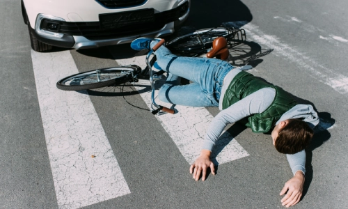 A cyclist lies injured on a crosswalk after a collision with a car, with the bicycle tangled under the vehicle’s front bumper highlighting the severity of bicycle accidents.