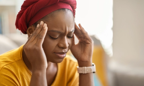 A woman sits indoors with her eyes closed and hands pressed to her temples, visibly in pain representing the physical and emotional toll of a traumatic brain injury.