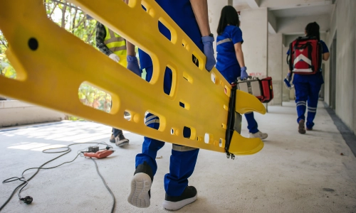 Emergency responders in blue uniforms carry a yellow stretcher through a construction site, indicating a workplace accident or injury on-site.