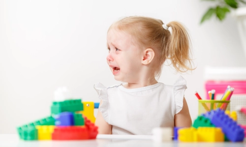 A young child with blonde hair cries at a table surrounded by colorful toy blocks and pencils, suggesting distress or possible injury at a daycare setting.