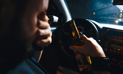 A person inside a car at night holding a beer bottle behind the wheel, with their other hand covering part of their face, depicting the danger and irresponsibility of drunk driving.