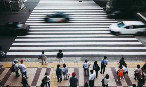 A crowd of pedestrians waits at a crosswalk while cars speed by, illustrating the potential danger of pedestrian accidents in busy urban areas.