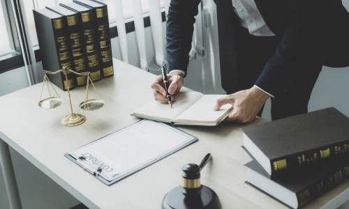 A lawyer in a suit writes in a notebook at a desk with legal books, a contract, a gavel, and the scales of justice, symbolizing a premises liability case review.