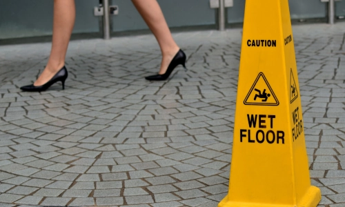 A woman in high heels walking past a bright yellow "Caution: Wet Floor" sign on a tiled surface, illustrating a potential slip and fall accident hazard in a public or commercial area.