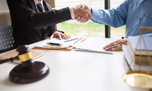 A lawyer and a client shake hands over a desk covered with legal documents, a gavel, and scales of justice representing an agreement or settlement in an Uber accident case.