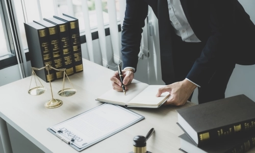 A lawyer stands at a modern desk writing in a notebook, surrounded by law books, a gavel, scales of justice, and a contract symbolizing the work of a premises liability lawyer handling property-related injury cases.