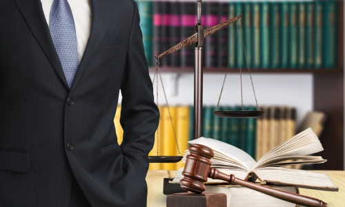 A Gainesville Lyft Accident Lawyer standing up while having one hand in his pants pocket while a law book and hammer and gavel are behind him.