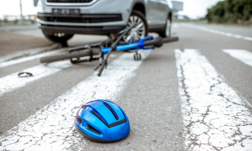 A blue bicycle helmet lies on a cracked crosswalk in front of a fallen bike and a silver SUV, indicating a bicycle accident scene.