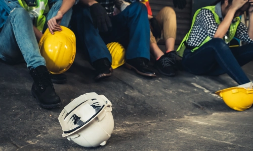 A group of construction workers sitting on the ground looking distressed, with safety helmets scattered nearby, symbolizing a workplace accident.