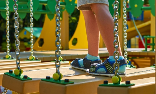 Close-up of a young child’s legs and feet walking on a suspended wooden bridge at a colorful playground, highlighting potential daycare safety hazards.