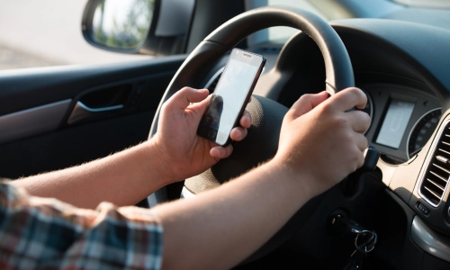 Close-up of a driver holding a smartphone while gripping the steering wheel with the other hand, inside a moving car—depicting distracted driving behavior.