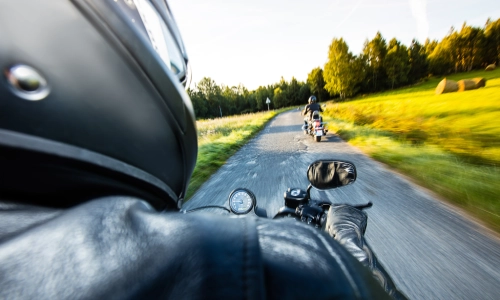 A dynamic view from behind a motorcyclist riding on a country road, with another biker ahead, surrounded by trees and open fields—capturing the speed, risk, and vulnerability associated with motorcycle travel.