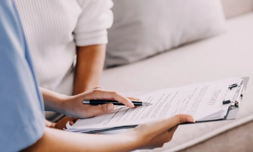 A nurse in scrubs holding a clipboard discusses documents with a seated elderly woman highlighting potential concerns related to nursing home abuse and patient care oversight.
