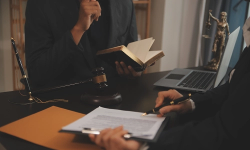 Two attorneys in suits review legal documents at a desk with a gavel, law book, Lady Justice statue, and open laptop, representing personal injury legal consultation.